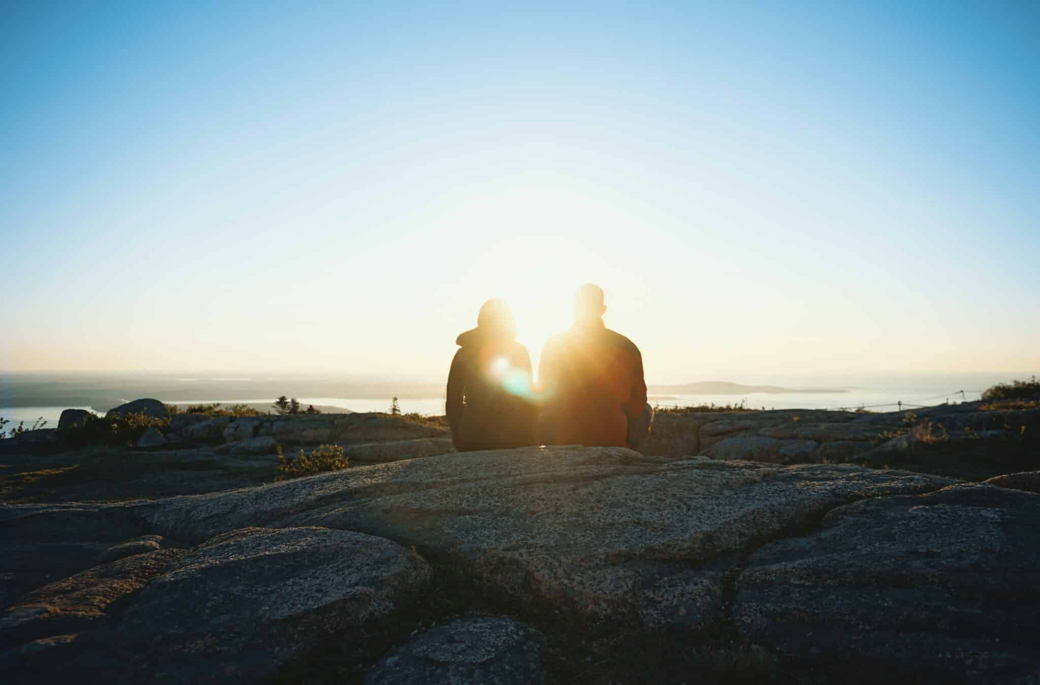 a younger couple sat on a rocky landscape looking out at the sunset